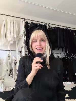 A woman wearing a black hoodie is holding a microphone and smiling at the camera. Behind her are racks of clothing. The composition is a medium shot, focusing on the woman from the chest up.