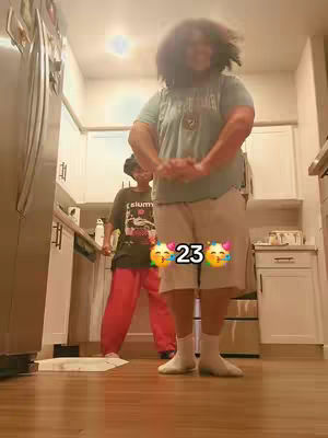 The image shows two people in a kitchen setting. A person with curly hair stands in the foreground, wearing a light blue t-shirt, khaki shorts, and white socks. They are positioned with their hands in front of them. In the background, another person is visible, wearing a dark shirt and red pants. The kitchen has white cabinets and silver appliances. The number "23" is overlaid with two emoji face stickers.