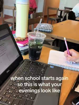 A laptop sits open on a wooden table, with a full drink in a Panera Bread cup to the right. A hand with a pink pen is writing in a spiral notebook. In the background are other tables and chairs, partially out of focus.