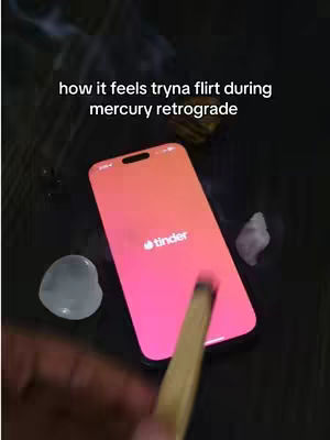 The image shows a phone with the Tinder logo on the screen, and a burning stick near it. The phone is sitting on a dark surface with crystals and hazy smoke around.