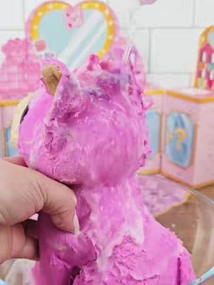 A product is being cleaned and revealed. The product appears to be a stuffed animal toy. The toy is pink. The cleaning action involves the use of a white soapy substance. In the background are pink background and decorative elements, including a heart-shaped mirror.