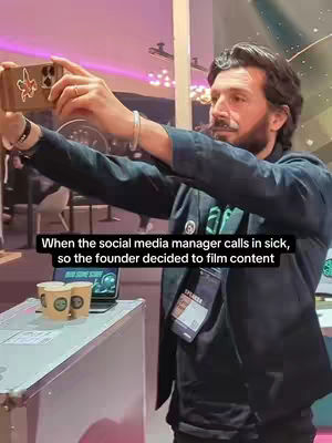 A man wearing a dark jacket takes a selfie with a smartphone in a conference setting. Text overlayed at the bottom reads: 'When the social media manager calls in sick, so the founder decided to film content.' The man has a beard and is wearing a name tag that says 'SPEAKER'. In the foreground are two coffee cups and a laptop.