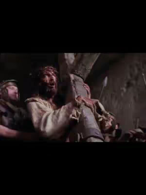 A low-angle shot from a film, featuring Jesus carrying a heavy wooden cross. He is mid-stride, visibly strained and wounded with a crown of thorns and blood on his face. A guard is visible behind him. The image is dark, emphasizing the dramatic nature of the scene.