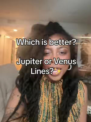 A selfie-style video cover with a woman with dark curly hair, posing indoors. Text overlay reads "Which is better? Jupiter or Venus Lines?" The woman smiles, wearing hoop earrings, her eyes focused on the camera. The background is softly blurred, suggesting an interior setting.