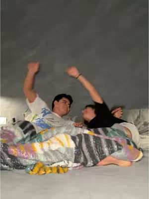 A couple is lying in bed, captured from a low angle with an overcast lighting that suggests a casual setting or moment of relaxation and comfort. The individuals are mostly covered by a colorful blanket with a patchwork design. The grey backdrop shows an even, solid color, which provides a neutral background. One person's arm is raised with a bracelet and the other is also raising an arm. The composition is framed to focus on the two people and their environment.