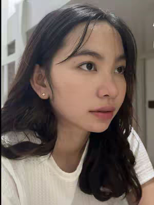 A close-up shot of a young woman with long dark wavy hair, fair skin, and a slight smile. She wears a white shirt and has a small earring in her ear. The background is a blurred interior space. The lighting is soft and natural.