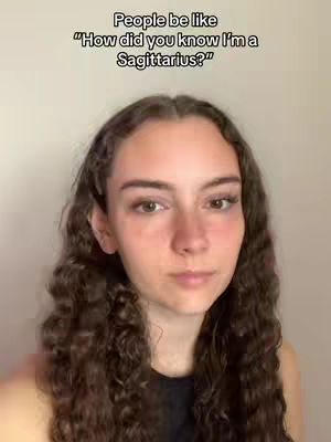 A young woman with long, wavy brown hair looks directly at the camera. The text overlay is in a white rectangular box, and reads 'People be like "How did you know I'm a Sagittarius?"'. The background is a plain, light wall.