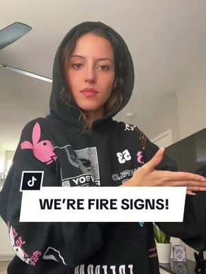A woman wearing a black hoodie with a hood up, her face is in the center, in front of a light-colored wall. A white rectangle with the text "WE'RE FIRE SIGNS!" is placed below her, a TikTok logo is on the left bottom corner.