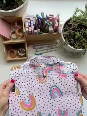 The cover image shows a well-lit, overhead shot showcasing a white surface filled with various stationery and an open bible bag. The bible bag, centrally positioned, is adorned with a pattern of rainbow arcs and polka dots. To the upper left, a wooden organizer holds colorful pens and markers. Next to the bag, a small wooden caddy displays washi tape rolls. Additional items like pens, pencils, and small green plants in white pots complete the composition.