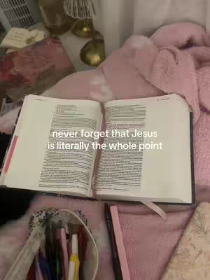 An open bible with text in an interior space. There are other objects, such as pens, a book, and a lamp in the frame.