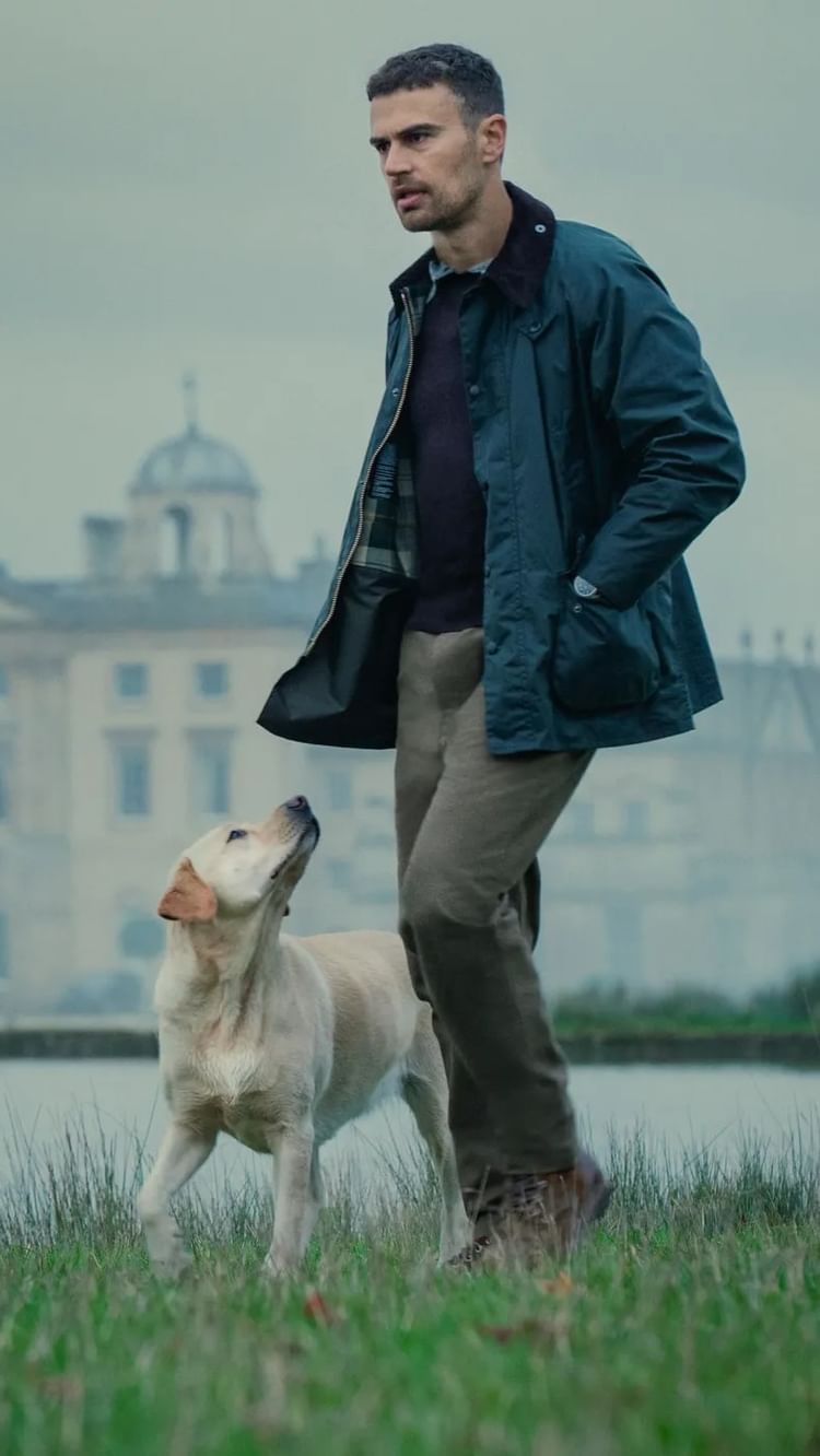 A man in a dark green jacket and brown pants walks in a field with a dog, a cream-colored Labrador, looking up at him. A building can be seen in the background. The overall composition is naturalistic, with muted colors and a focus on the relationship between the man and the dog.