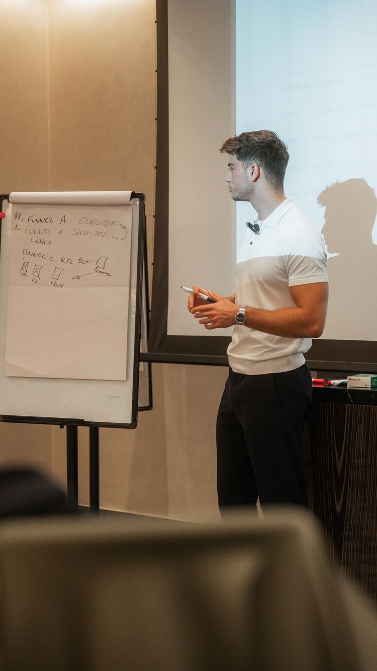 A man in a white and grey polo shirt is presenting in front of a whiteboard, with a blurred background of a conference room. He stands to the side, and the whiteboard has diagrams and text on it.