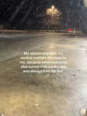 The image shows a blurry scene of what appears to be a wet outdoor environment during a snowfall. The ground is covered either in water or ice. The central area is blurred with visible raindrops and falling snow. In the background, light from a street lamp illuminates the scene, casting a soft glow on the surrounding area.