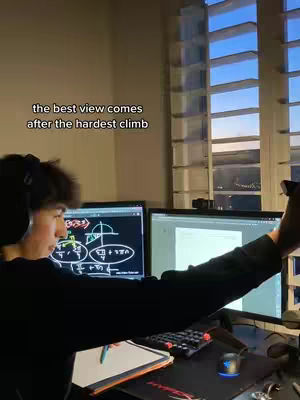 A young man with dark hair sits at a desk with two computer monitors, a keyboard, a mouse, and a tablet. One monitor on the left displays a diagram with lines and shapes, while the right screen is mostly blank. The man is holding a phone or another device in his right hand, angled toward the camera. The background includes a window with a view of buildings lit by what appears to be evening light.