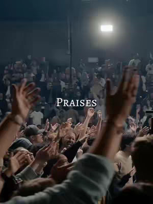 A low-angle shot of a large crowd in a concert setting, with many hands raised in the air. The foreground is filled with hands, and the background includes blurred figures and stage lighting. The word PRAISES is superimposed in the center of the image, slightly above the heads of the crowd.