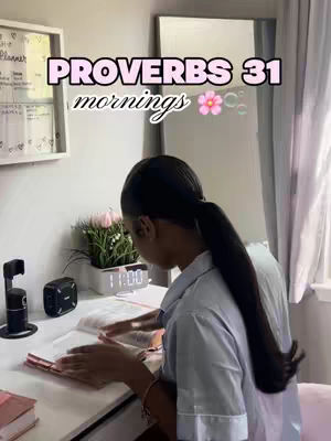 A woman is sitting at a desk, leaning over a book. The desk is white and cluttered with various items, including a lamp, a speaker, and a pink flower arrangement. The woman has a ponytail and is wearing a light-colored shirt. Above her, a text overlay reads "PROVERBS 31 mornings" with a flower graphic.