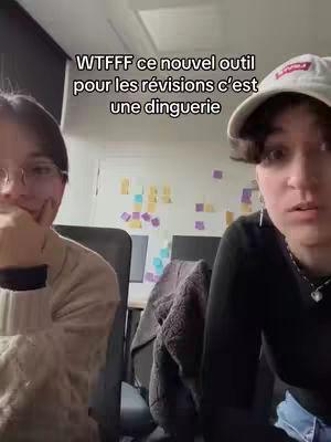 Two people are seated. The focus is on the two people, a wall covered in sticky notes behind them. The person on the right has a white baseball cap. The person on the left is wearing glasses and has their hand on their chin, as if thinking. The one on the right is turned toward the camera and speaking.