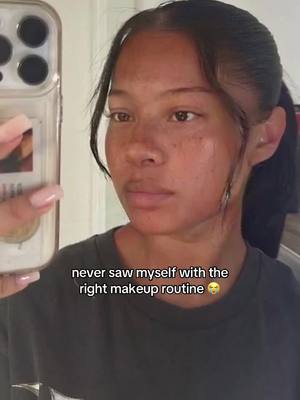 A young woman is holding a phone in front of her face to record a selfie-style video. She is wearing a dark gray t-shirt. The image has a medium shot, focusing on her face and upper body. The background is blurred, suggesting an indoor setting. Text is overlaid at the bottom: 'never saw myself with the right makeup routine' with a smiley face.