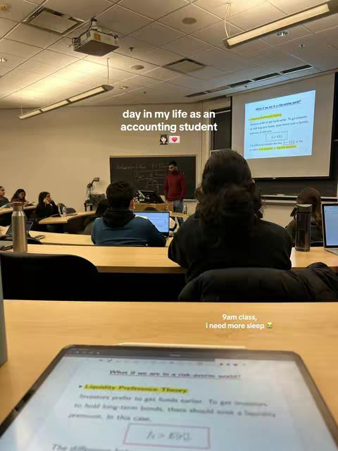 A photo taken from the perspective of someone sitting in a classroom. The focal point is a laptop on a wooden desk, displaying a page of notes titled 'Liquidity Preference Theory,' highlighted with yellow and pink markers. In the background, other students are present in the classroom. A teacher stands at the front near a projector screen with a presentation visible. Above, the words 'day in my life as an accounting student' are written in the image, along with additional text: '9am class, i need more sleep'.
