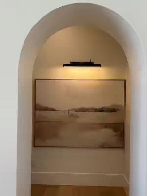 An abstract painting hangs within an arched doorway. Above the painting, a small black light illuminates the artwork. The artwork itself features a landscape painting with neutral earth tones.