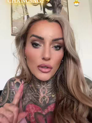 A close-up shot of a woman with blonde hair, heavy makeup, and multiple tattoos, pointing a finger towards the camera. The background is a blurred interior with a painting.