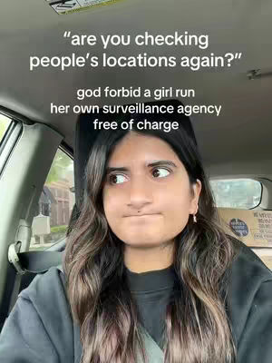 A woman is sitting in the driver's seat of a car, looking to her left with a slightly annoyed expression. The image is composed of the woman's face and upper body. Overlaid text says 'are you checking people's locations again?' and 'god forbid a girl run her own surveillance agency free of charge'. The lighting is natural appearing from outside the vehicle.
