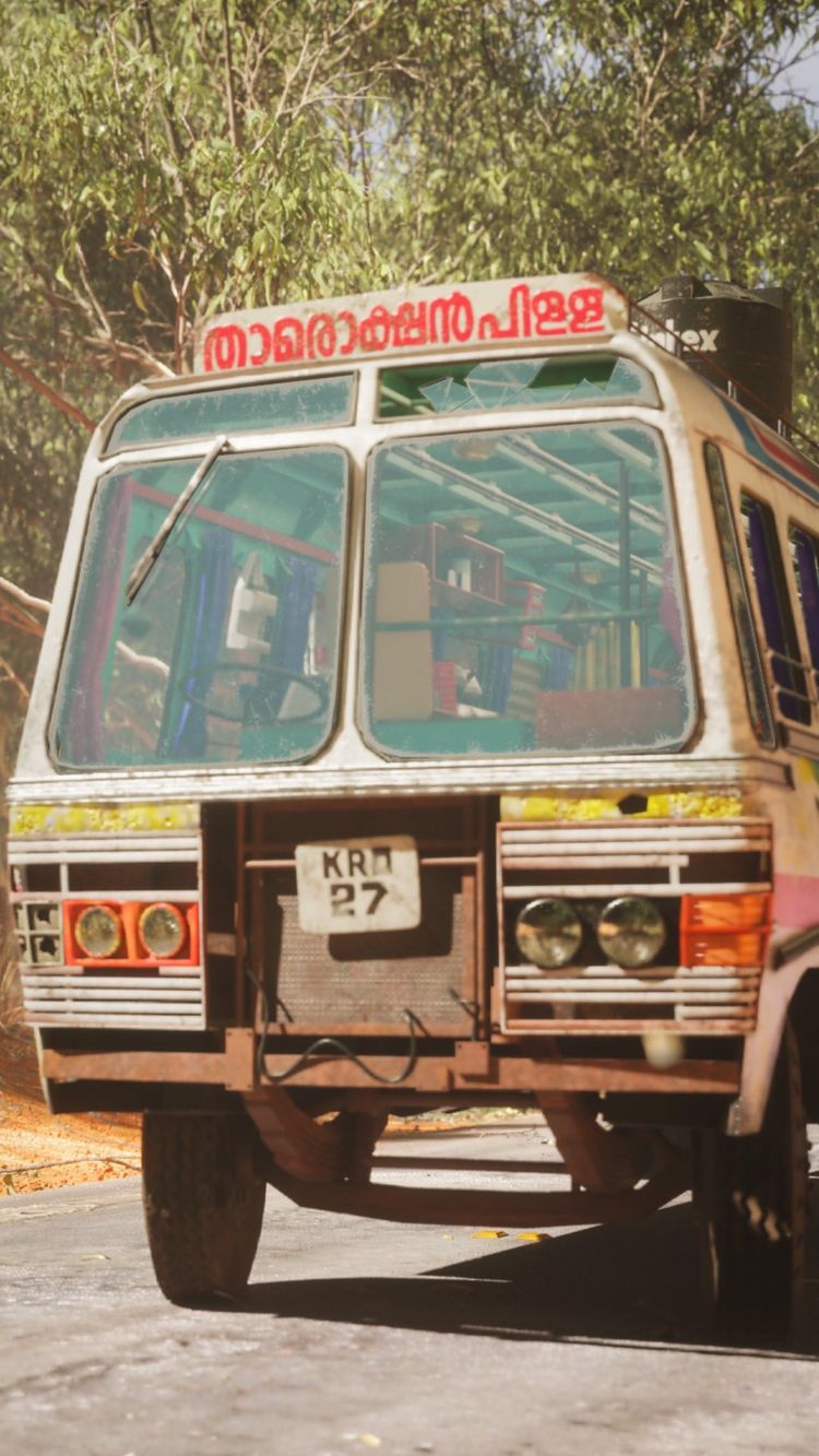 The image showcases a well-detailed, slightly worn, vintage-style bus from a low angle. The bus is primarily cream-colored with red and pink accents. The front of the bus is the focal point, with large rectangular windows offering a glimpse into the interior, likely containing seating or cargo. A sign with text in a non-English script is visible above the front windshield. The bus has a distinctive number plate and is set against a backdrop of foliage. The lighting is bright.
