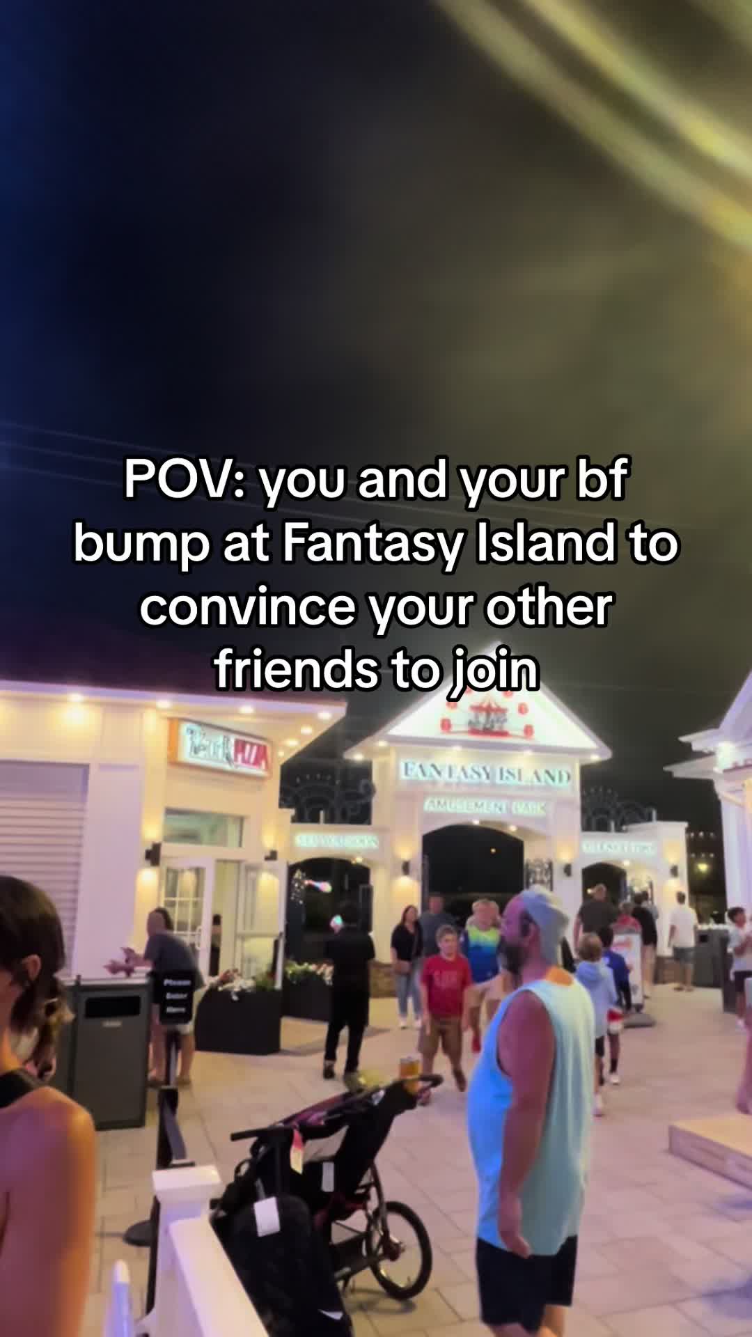 An outdoor shot at night at Fantasy Island amusement park. In the foreground, a person in a blue shirt and black shorts is standing, facing away from the camera. A stroller with a black storage bag is right beside him. In the midground, a group of other people can be seen walking. The background contains the entrance of Fantasy Island, with signage. The sky is dark, and the area is lit with lights.
