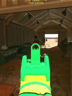 The first-person perspective shows the view down the sight of a gun in a video game, inside a building or structure. The bright green and yellow sight is the focal point, with a blurred background showing players and the game environment.
