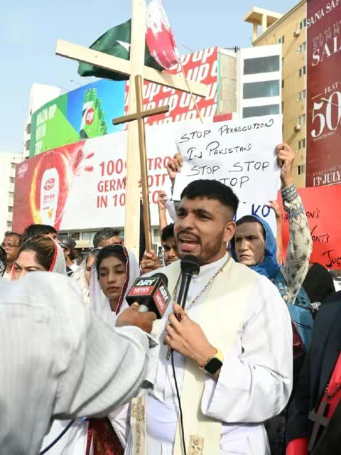 The cover image shows a man in a white clerical shirt speaking into a microphone, likely at a protest. Behind him, a crowd of people is visible. Some hold signs with text, and a large wooden cross stands prominently. Flags of Pakistan and a related flag are visible. The setting appears to be an outdoor public space, with building signs in the background.