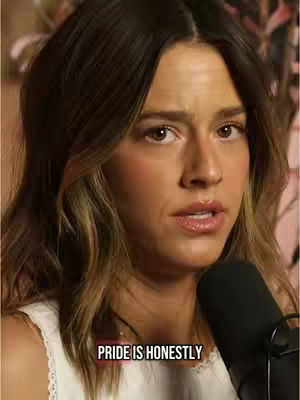 A close-up shot features a woman with brown hair and a neutral expression. The background is blurred, but hints of a floral pattern are visible. A microphone is positioned in the bottom right corner, and text overlayed at the bottom, reading "PRIDE IS HONESTLY".