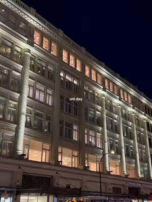 A nighttime shot of a large multi-story building with numerous lit windows. The building has classical architectural features like columns. The sky is dark blue.