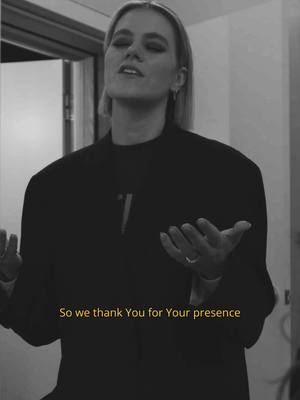 A black and white photo of a woman in a dark top and light hair singing. She is in what looks like a building, with her eyes closed and her hands open. Text appears at the bottom, also in yellow.