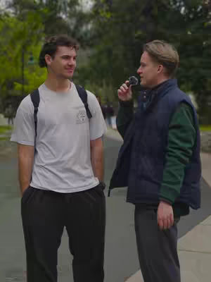 Two men stand outside, one seemingly interviewing the other. The interviewer holds a microphone to his mouth, while the interviewee stands with his arms relaxed at his sides. They're in what appears to be a park or outdoor area, with trees in the background.
