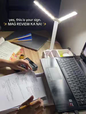 A well-lit desk with a laptop, books, and study materials. The primary focus is a white desk lamp casting light, while there are also papers and a calculator open on the desk, suggesting a study session is in progress.