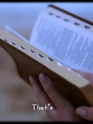 The image shows a person holding an open book, presumably a Bible, with their hands in the foreground. The book is open to a page with visible text, and the edges of the book are brown with cut-out tabs. The background is blurred, implying a soft focus on the book. In the bottom section, right corner, the text "That's" is overlaid.