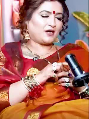 The image shows a woman of Indian descent. She has dark hair styled with a bun. She has a bindi on her forehead and is wearing multiple rings and bracelets. She is holding a microphone and there is a leafy green object in the background. The woman is speaking and an orange and red saree covers her body.