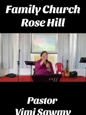 The image is a vertical video still. The top third has the text overlayed 'Family Church Rose Hill' in white, bold, sans-serif font on a black background. Below that, there's a live shot of a woman speaking into a microphone, in a church setting. The background consists of a projection screen, curtains, musical equipment, and some guitar. Under the live shot, there is another black background, with the text 'Pastor Vimi Sawmy' in white, bold, sans-serif font.