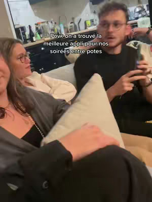 A candid shot of three people seated on a couch in a home setting. The focus is on a person in the center, holding a phone, who seems to be the focal point, and the two others are casually displayed. The lighting is warm, suggesting an indoor setting.