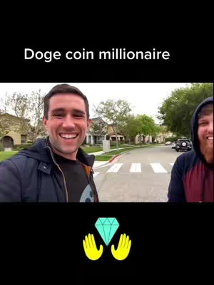 A video cover image featuring two men smiling and standing outdoors on a neighborhood street. The background includes houses, trees, and a road marked with pedestrian crosswalk lines. Overlaid at the bottom of the image is a graphic of a green diamond with yellow hands on each side.