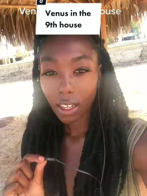 A medium shot shows a woman speaking to the camera with a text overlay. She has long, black braided hair, and is wearing a green top. She has on eyelash extensions and a nose ring. The background is a blurry mix of sand, plants, and sunlight. The text overlay says: Venus in the 9th house.