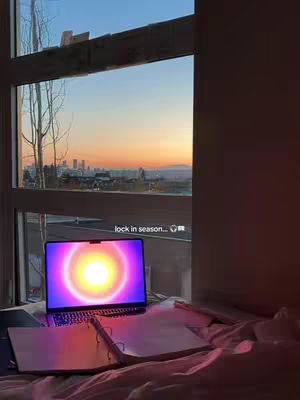 A photo shows a well-lit room with a large window overlooking a cityscape at sunset. A laptop with a glowing screen and open notebook is set up on a desk near the window. The setup suggests a study or work environment with a view.