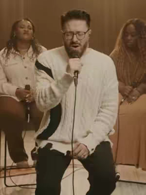 A man with glasses singing into a microphone is centered in the frame, wearing a white sweater with black accents and distressed details. Two women are seated on either side of him in the background, one with their eyes closed and the other looking down, all on a stage setting with warm lighting.