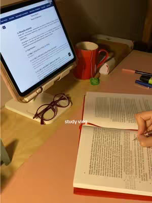 A desk setup for studying at night featuring a laptop displaying text, a red mug, and an open book with a hand writing.