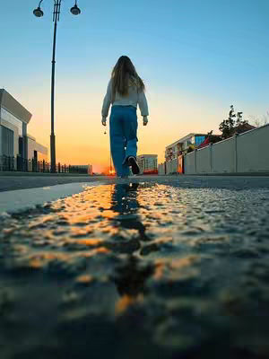 A low-angle shot of a person walking away down a road towards an orange sunset. The road surface is wet, creating reflections. A lamppost is on the left, and buildings are in the background. The sky is blue, transitioning to orange. The person is wearing light-colored clothing, adding contrast to the scene.