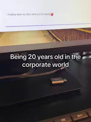 The image shows a computer monitor with a screen displaying text. Below the screen is a black surface with a cable and a small, gold-colored device. The text overlay reads 'Being 20 years old in the corporate world'. The overall composition is simple, with the focus on the screen and the accompanying text.