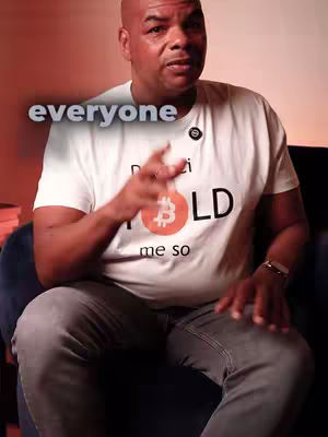 A man with a shaved head sits in a chair, wearing a white t-shirt with the Bitcoin logo on the front and the words "Davinci Hold me so" printed on it. Text overlay says "everyone". He is pointing towards the viewer. The background is blurred with a warm, orange-red glow.
