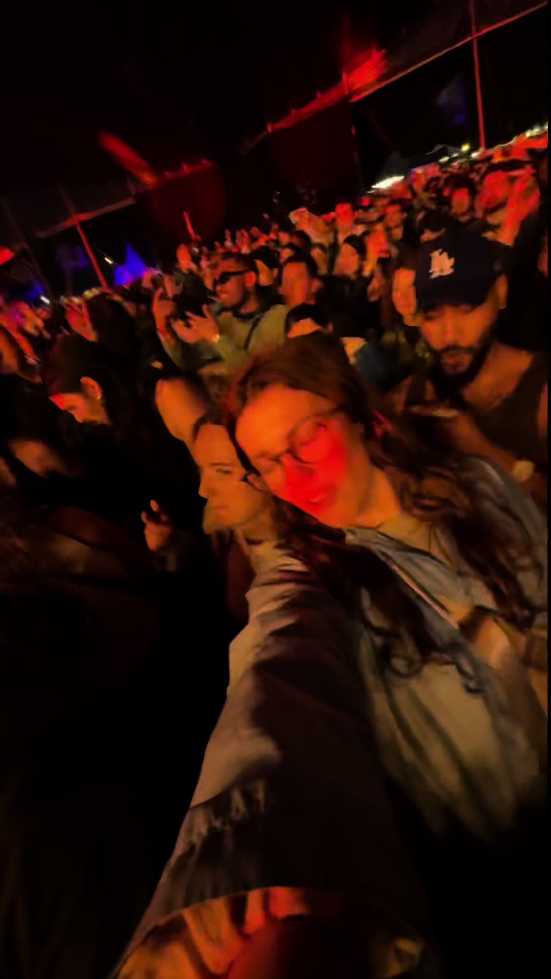 A selfie-style shot taken at a music festival. The foreground is dominated by a person with glasses and long hair, illuminated with red stage lighting. Behind them, a large crowd is visible, immersed in a concert with the stage out of view. The composition suggests a lively, nighttime atmosphere, with the flash of the camera adding brief illumination, capturing a sense of energy and shared experience among attendees.