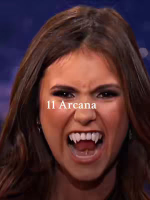 A close-up shot of a woman's face, mid-expression. She is baring her teeth, with prominent, elongated vampire teeth. The text "11 Arcana" is overlayed at the bottom center. The background is out of focus and a mixture of purple and blue.