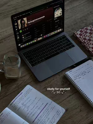 The image showcases a workspace setup with a laptop prominently displayed open on a wooden surface. The laptop screen shows what appears to be a music streaming service interface, alongside two open notebooks and a glass of water. The overall composition has a warm, inviting aesthetic.